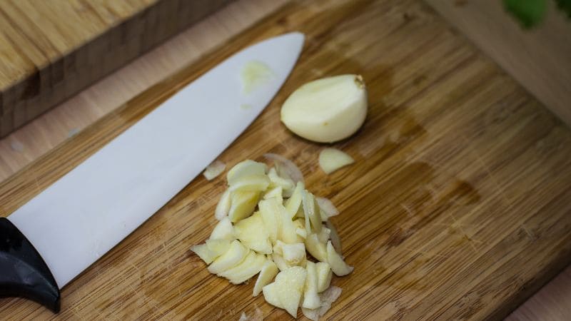 Ceramic knife with a black handle next to chopped garlic and a whole clove on a wooden cutting board