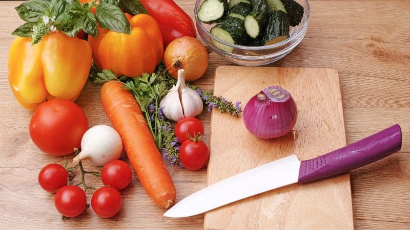 Ceramic knife with a purple handle on a wooden cutting board surrounded by fresh vegetables, including bell peppers, tomatoes, carrot, garlic, onion, and cucumbers in a bowl