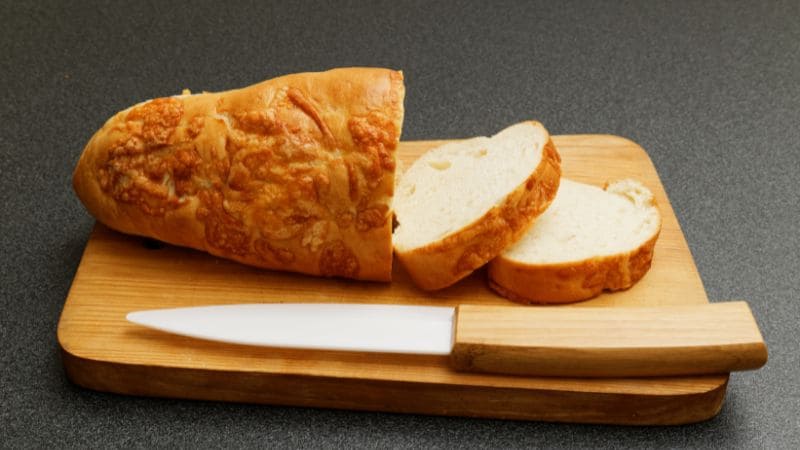 Ceramic knife with a wooden handle next to a loaf of cheese bread and two slices on a wooden cutting board