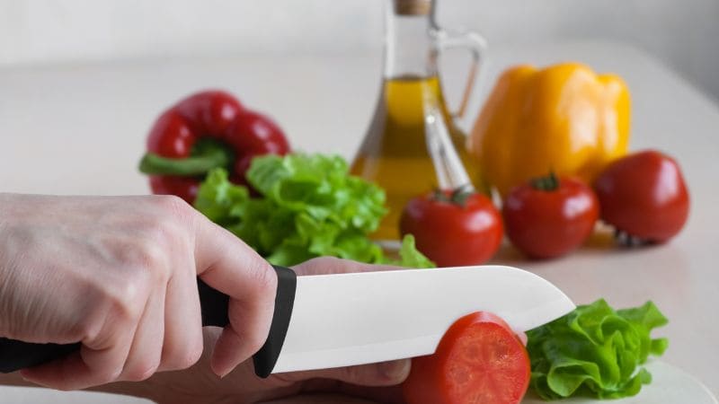 Person slicing a tomato with a ceramic knife on a kitchen counter, surrounded by fresh vegetables and a bottle of olive oil