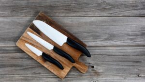 Set of three ceramic knives with black handles arranged on a rustic wooden cutting board over a weathered wood surface