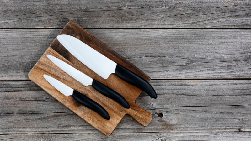 Set of three ceramic knives with black handles arranged on a rustic wooden cutting board over a weathered wood surface