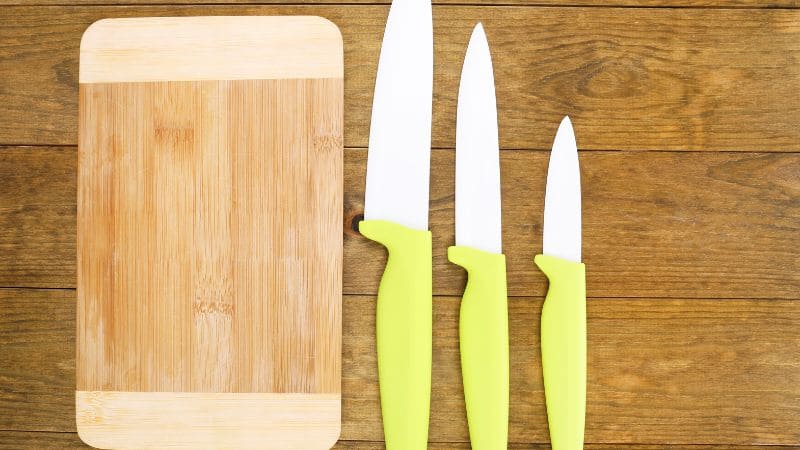 Three ceramic knives with lime green handles arranged next to a wooden cutting board on a rustic wooden surface